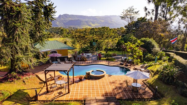 A swimming pool with a hot tub, surrounded by a patio with outdoor furniture, set against a backdrop of lush greenery and mountains. - Hotel Villa Florencia (Turrialba)