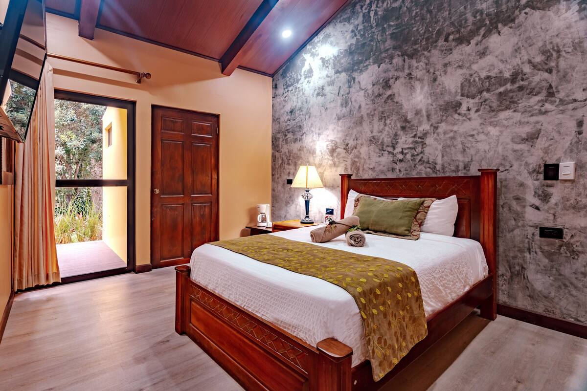 A bedroom with a wooden bed, a nightstand, a lamp, and a view of the outdoors through a door.