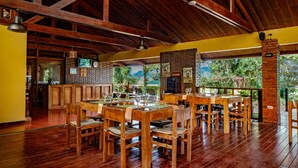 A restaurant with wooden tables and chairs, a brick fireplace, and a view of greenery outside. - Hotel Villa Florencia (Turrialba)