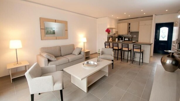 A modern living room with a sofa, coffee table, and two lamps. A kitchen with a dining area is visible in the background.