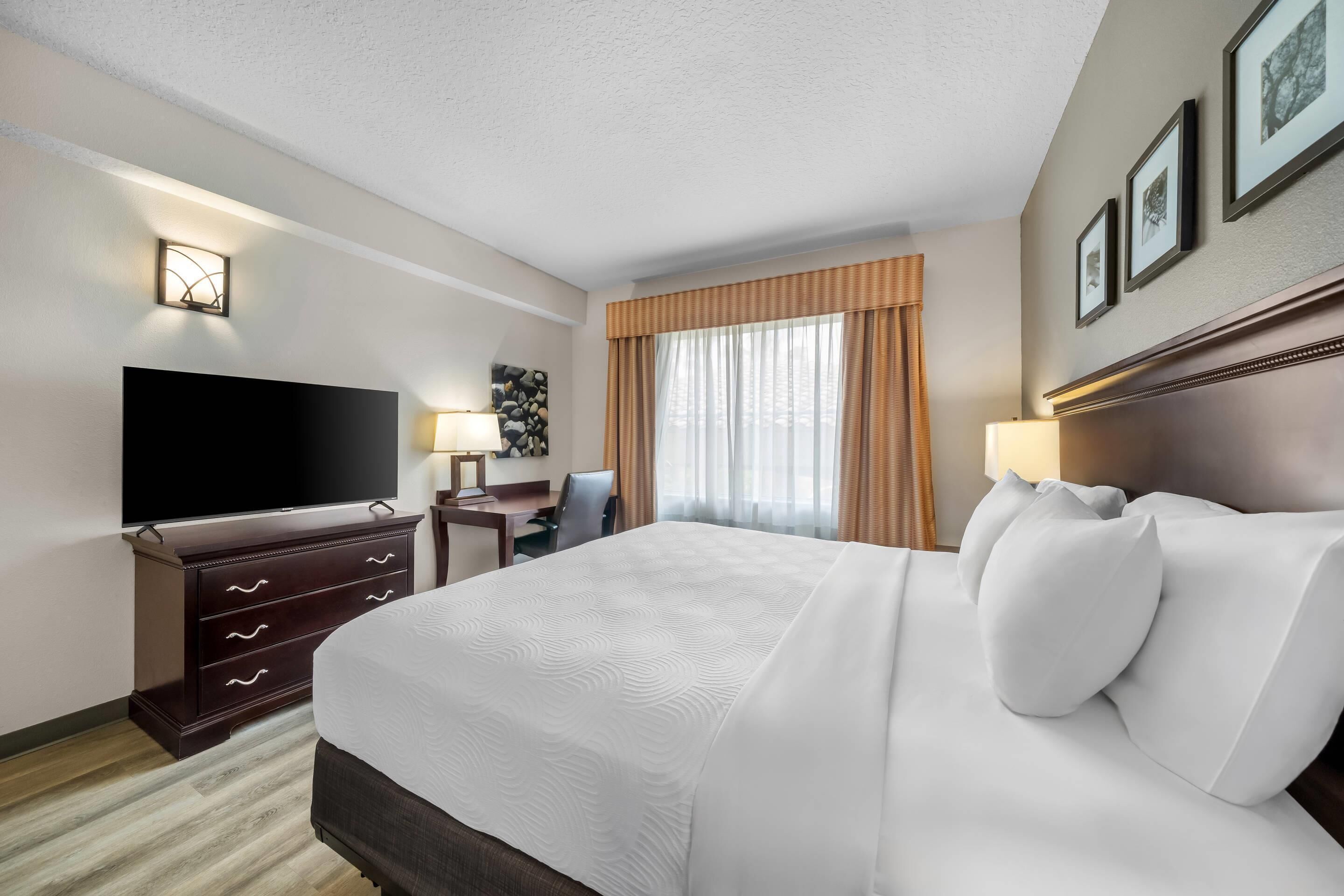 A hotel room with a large bed, a flat-screen TV, a desk with a chair, and a window with curtains.