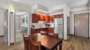 A modern kitchen with wooden cabinets, a stainless steel refrigerator, and a dining area with a wooden table and chairs. - Country Inn & Suites by Radisson, Port Orange-Daytona, FL (Port Orange)