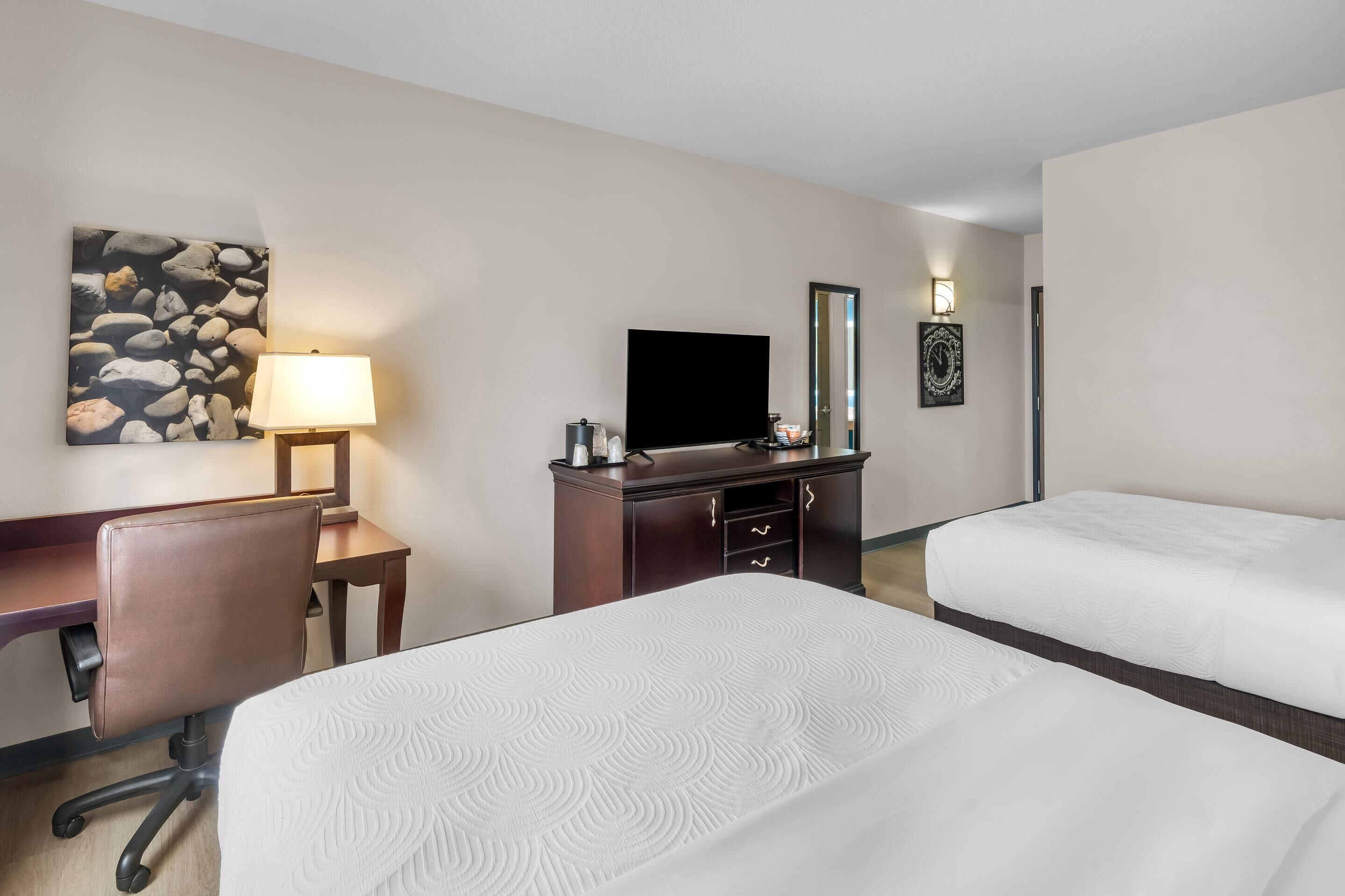 A hotel room with two beds, a desk, a chair, a TV, a lamp, and a wall art piece.