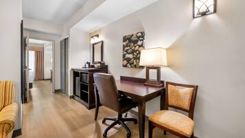 A hotel room with a desk, two chairs, a lamp, a painting on the wall, and a mirror.