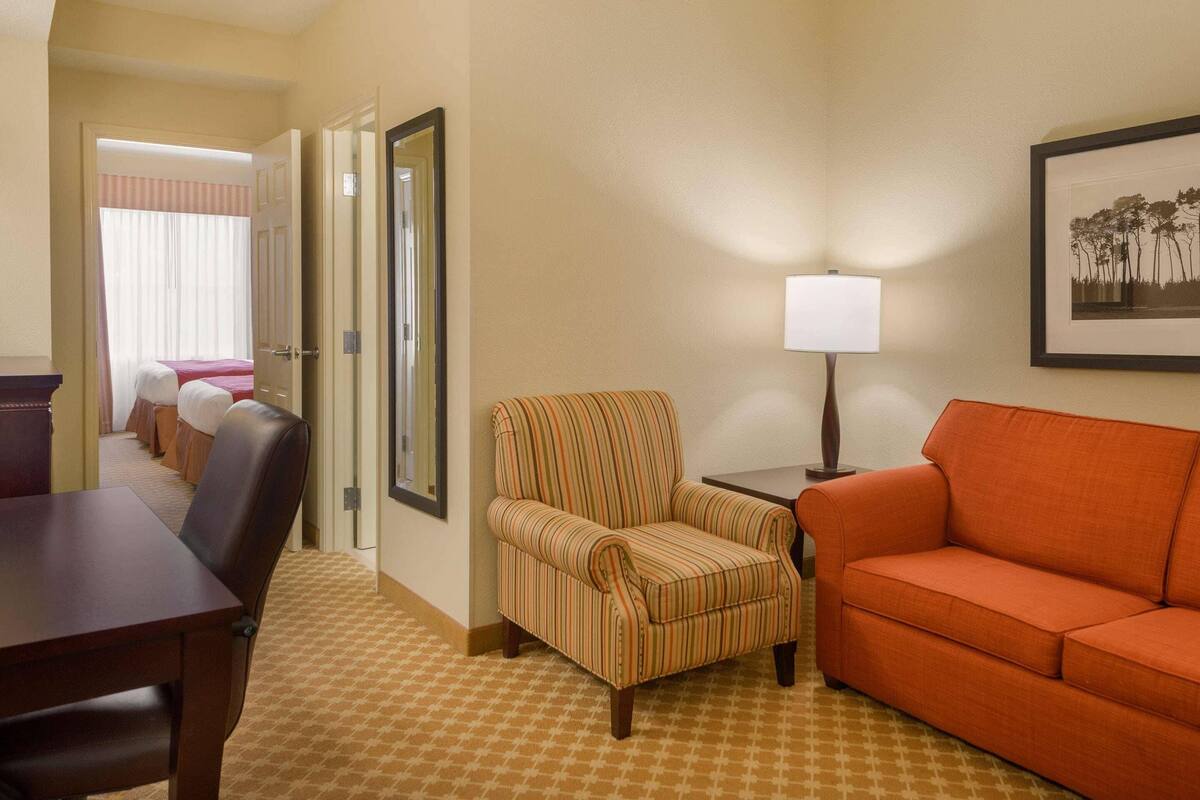 A hotel room with a dining table, striped armchair, orange sofa, and a lamp on a side table.
