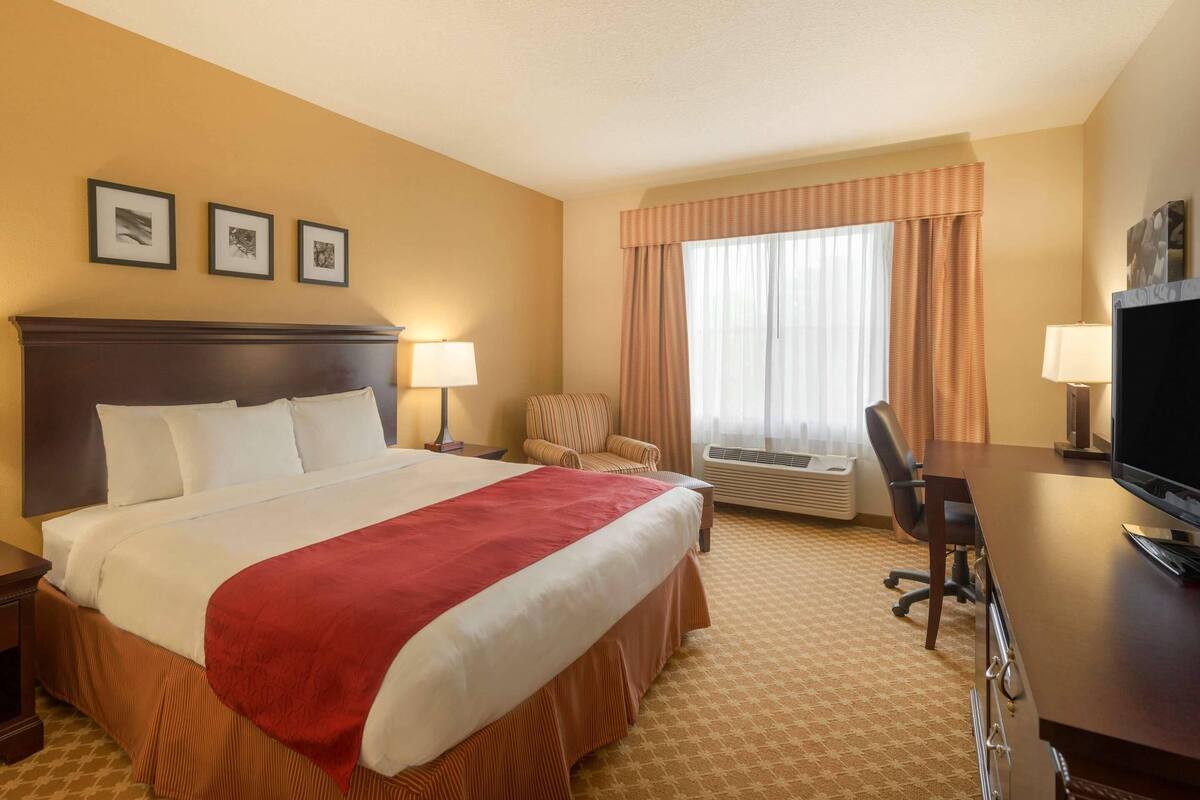 A hotel room with a large bed, a desk with a computer, an air conditioning unit, a chair, and framed pictures on the wall.