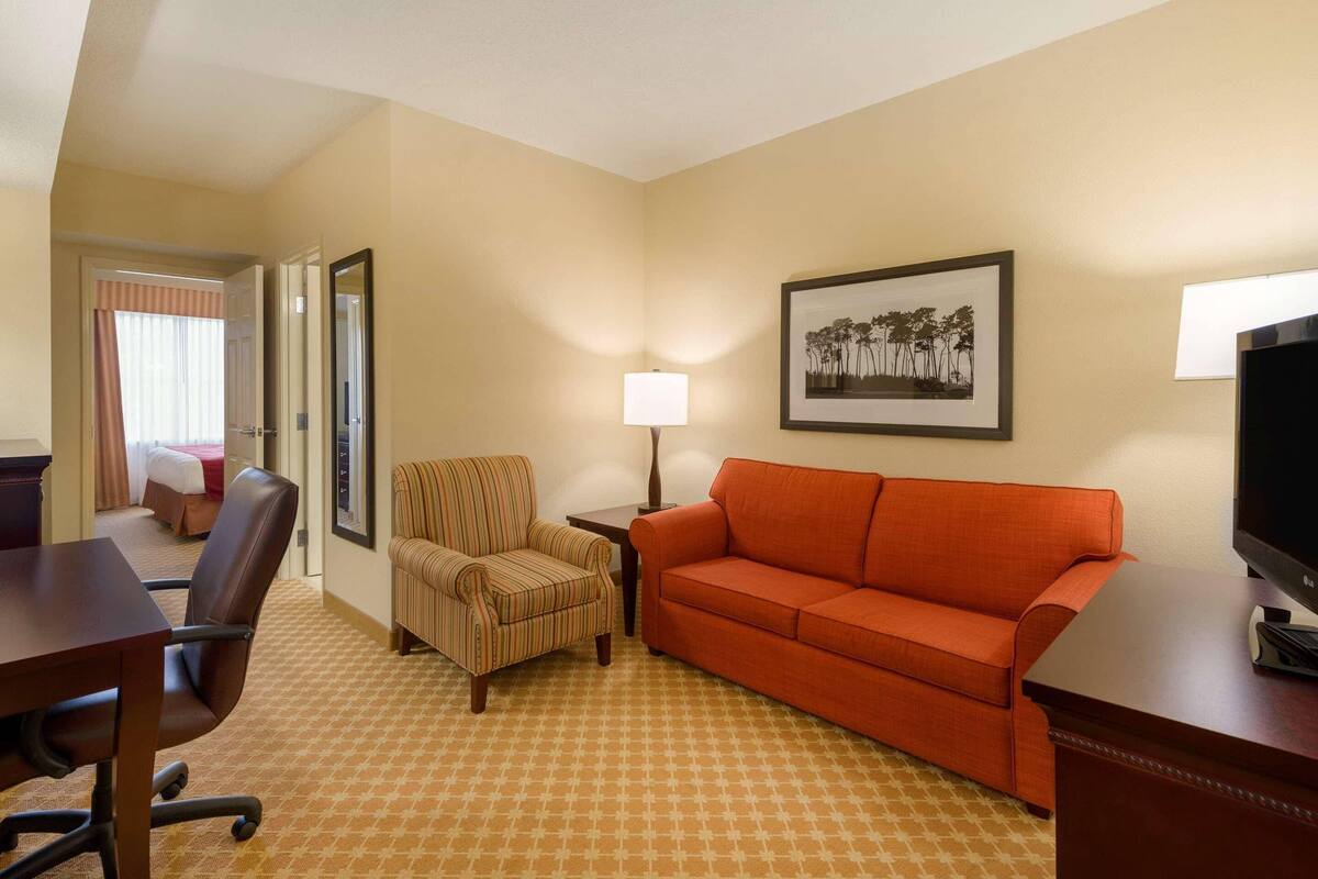 A hotel room with a desk, a striped armchair, an orange sofa, a lamp, a framed picture on the wall, and a TV on a stand.