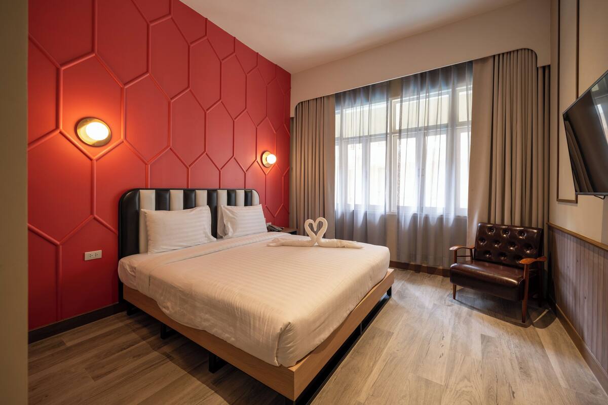 A modern hotel room with a large bed, a wooden floor, a red wall with hexagonal patterns, a large window with curtains, and a leather armchair.