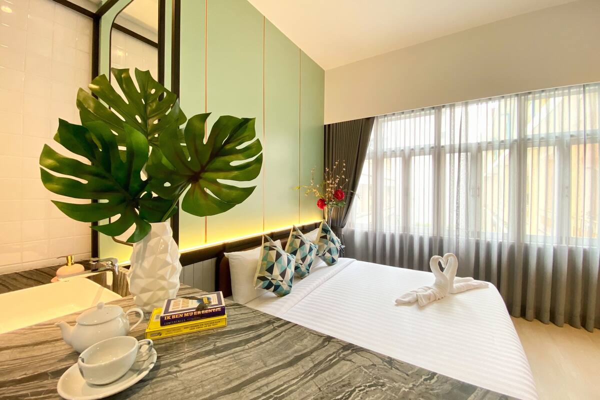 A hotel room with a bed, a large plant, a bedside table with a book, a teapot, and a cup.