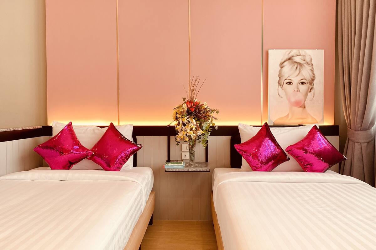 A hotel room with two beds, each with white bedding and pink star-shaped pillows. There is a small table with a vase of flowers between the beds.