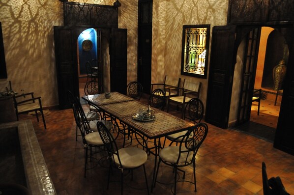Un espace repas avec une longue table et des chaises, entouré de murs ornés de motifs complexes et agrémenté d’une fenêtre à vitraux.