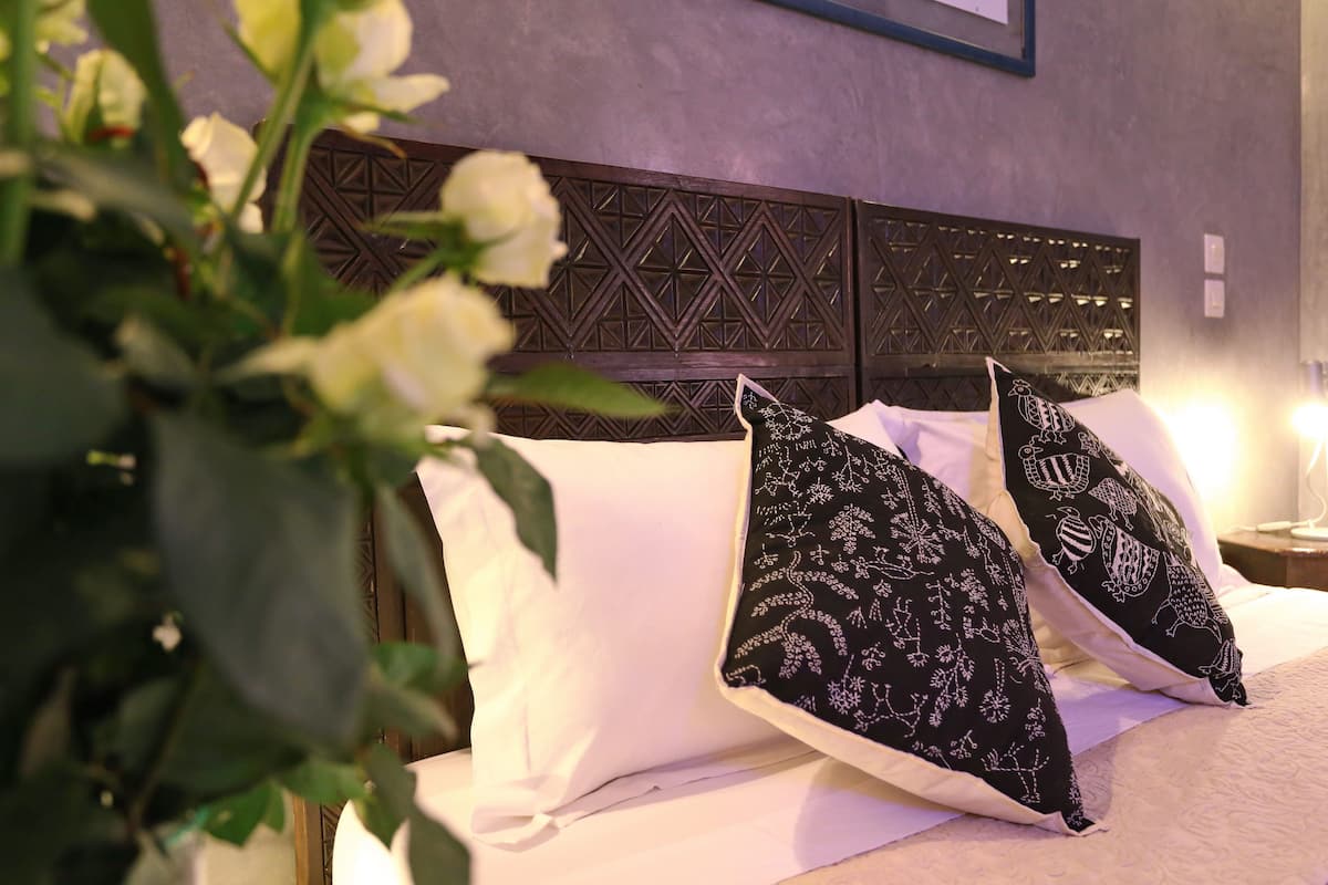 Una cama con sábanas blancas y almohadones decorativos, cabecera de madera con patrones geométricos, y una lámpara de pared.