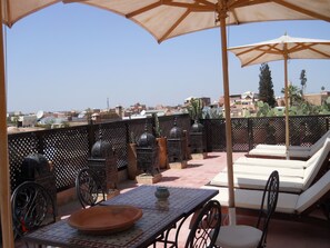 Une terrasse sur le toit, aménagée avec une table, des chaises, un transat et des éléments décoratifs, comme un cactus et des lanternes en métal.