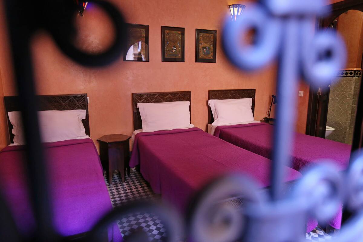 Habitación de hotel con tres camas, cada una con cubrecama morado y almohadas blancas. En la pared, sobre las camas, hay cuadros enmarcados.