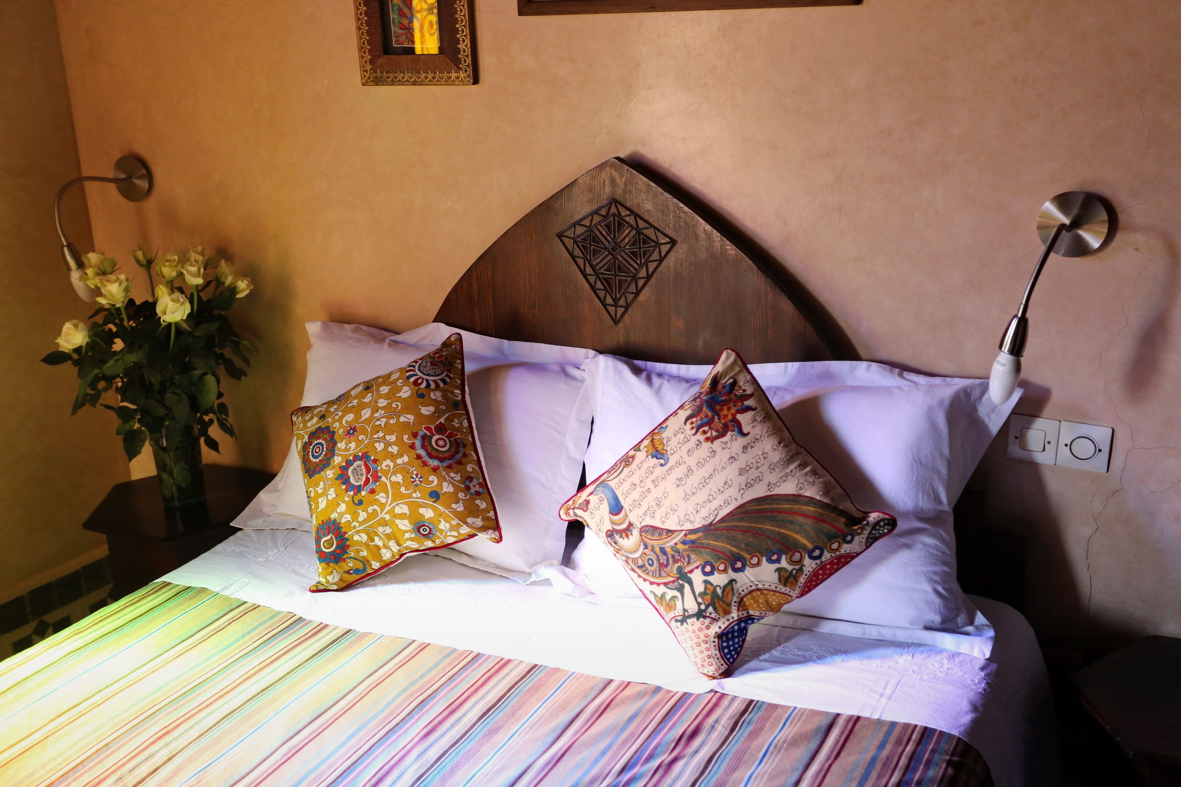 Un lit avec des coussins à motifs, un vase rempli de fleurs et un luminaire fixé au mur.