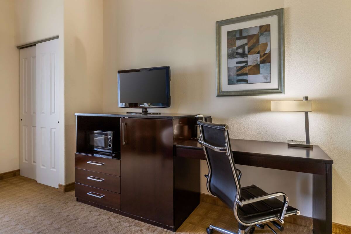 Habitación de hotel con un escritorio de madera oscura, un televisor de pantalla plana, una silla moderna, una lámpara y un cuadro abstracto enmarcado en la pared.