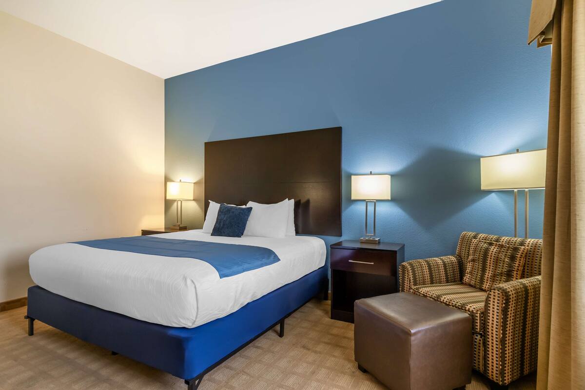 Una habitación de hotel con una cama grande, dos mesitas de noche con lámparas, un sillón estampado y un reposapiés marrón.