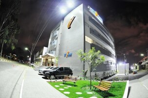Un hotel de diseño moderno, con un logotipo destacado en la fachada, un estacionamiento con coches y una pequeña zona ajardinada con bancos.