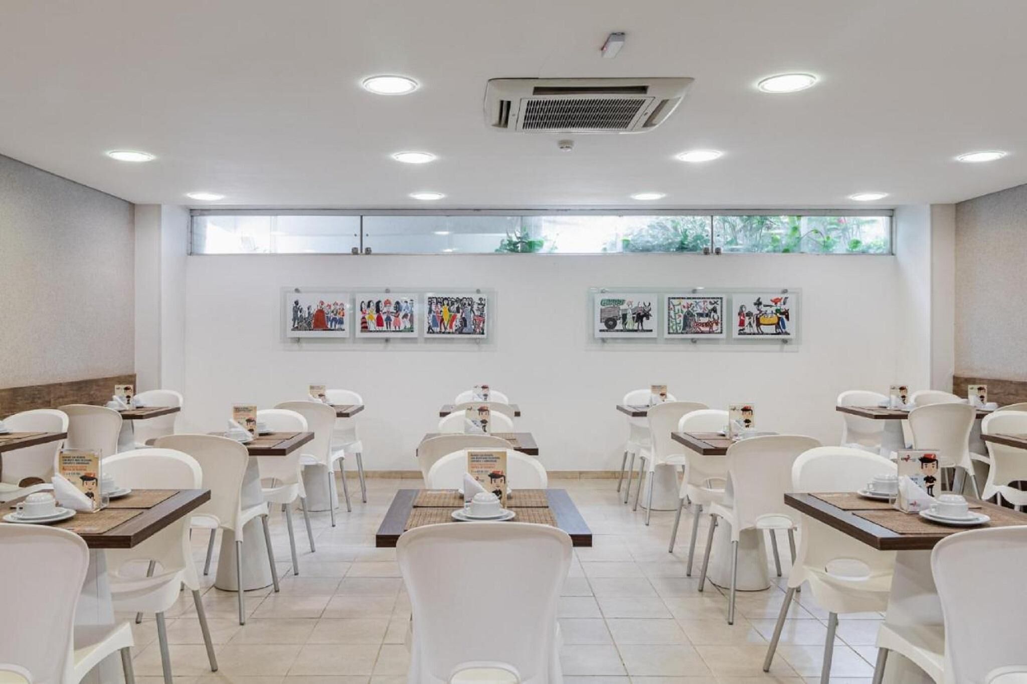Un comedor con sillas y mesas blancas, obras de arte en las paredes y un gran ventanal que proporciona luz natural.