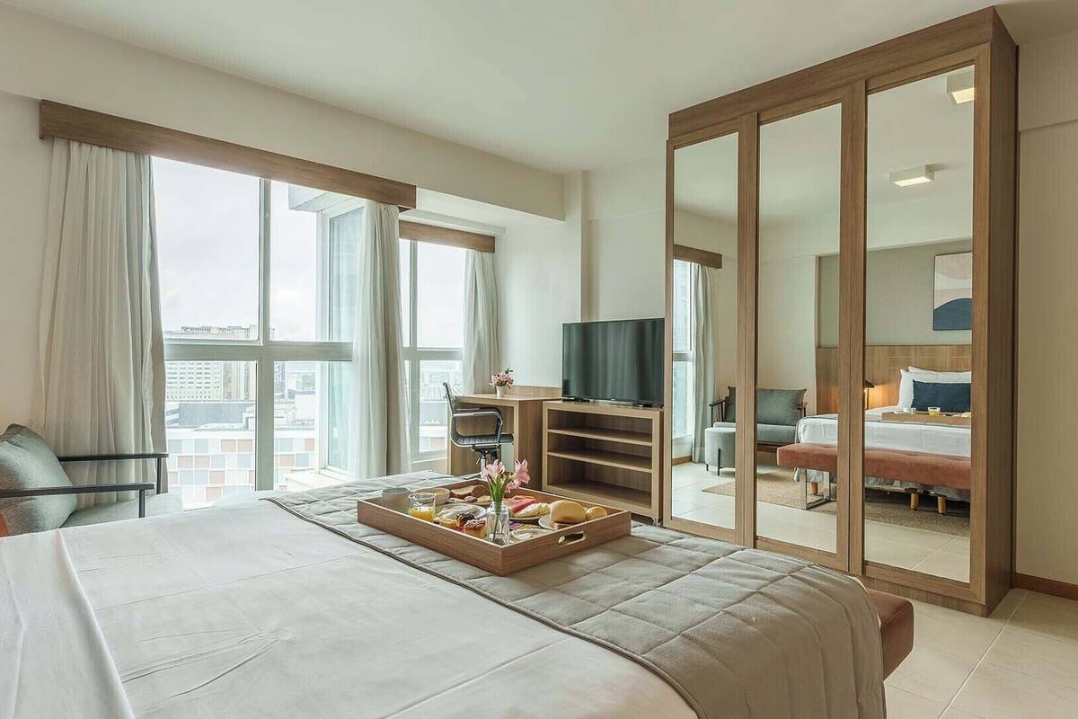 Quarto de hotel com uma cama grande, televisão, escrivaninha com cadeira, guarda-roupa e janela com cortinas.