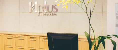 A reception desk with a computer monitor, a plant, and a sign that reads "Plus Hotelaria".