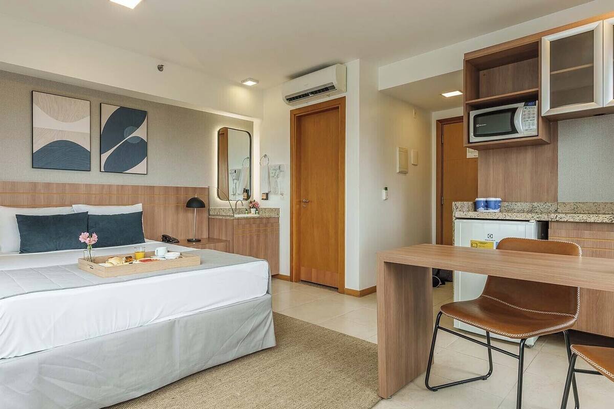 Quarto de hotel com cama, escrivaninha, cadeira, micro-ondas e pequena cozinha.