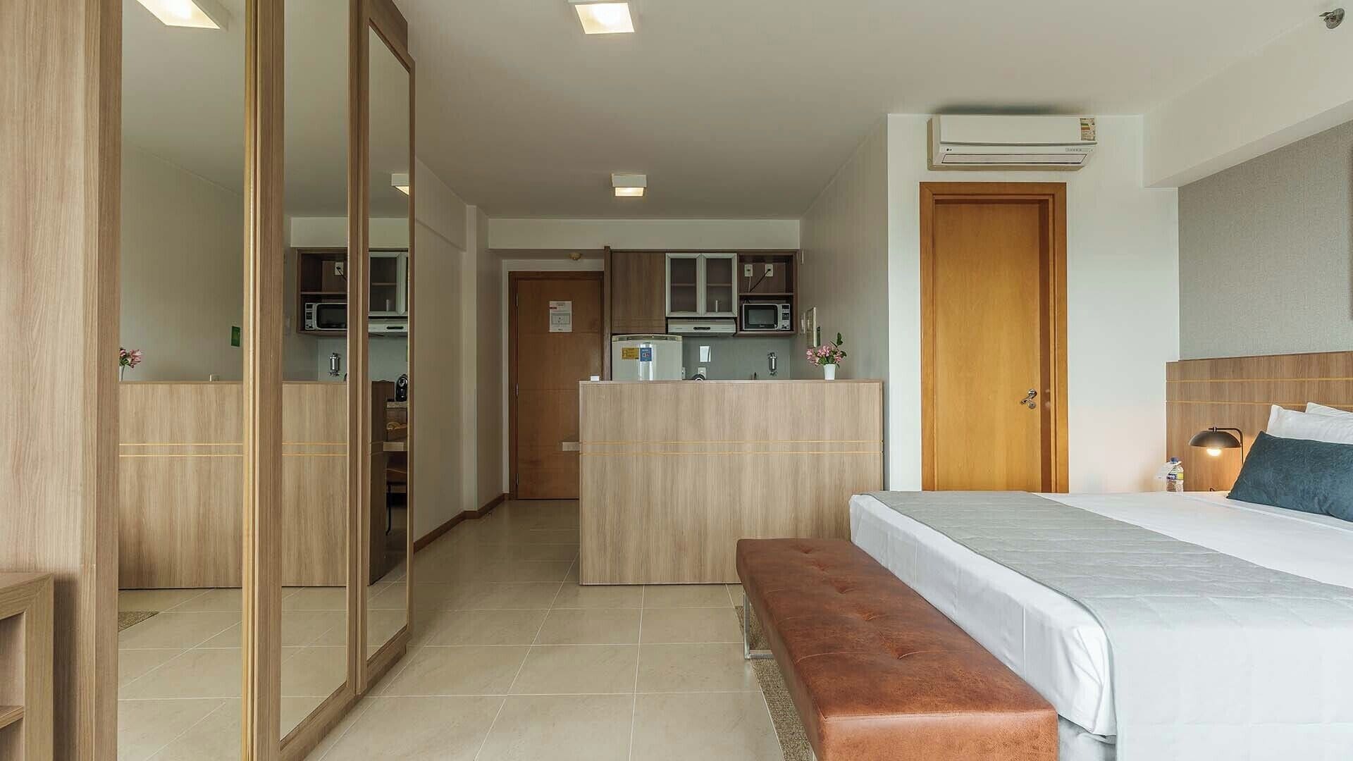 Una camera d'albergo con un letto, una panca, un armadio, uno specchio, angolo cottura e condizionatore d'aria.
