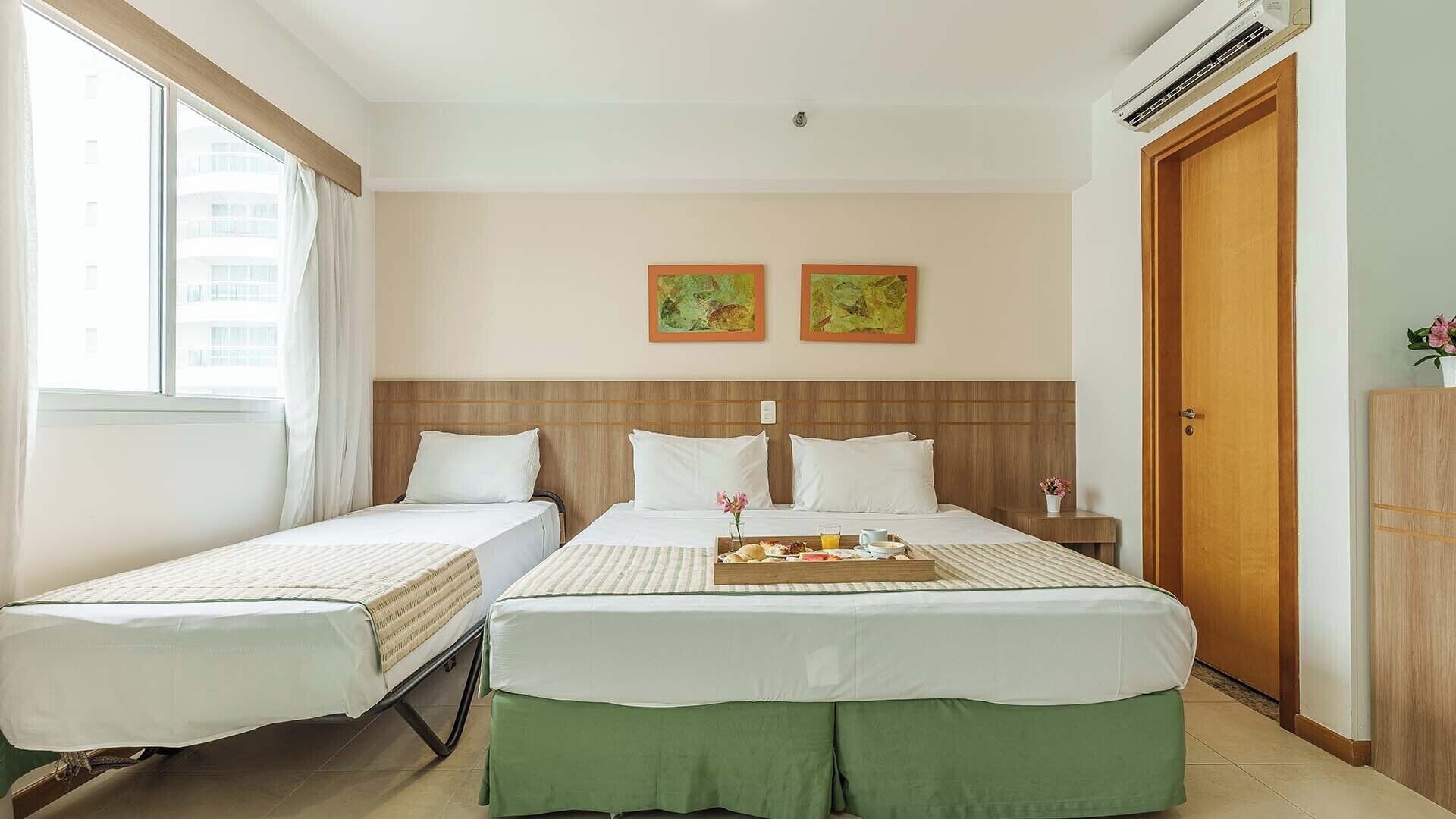A hotel room with two single beds, a larger bed, a wooden headboard, a nightstand, a tray with breakfast, and a window with curtains.