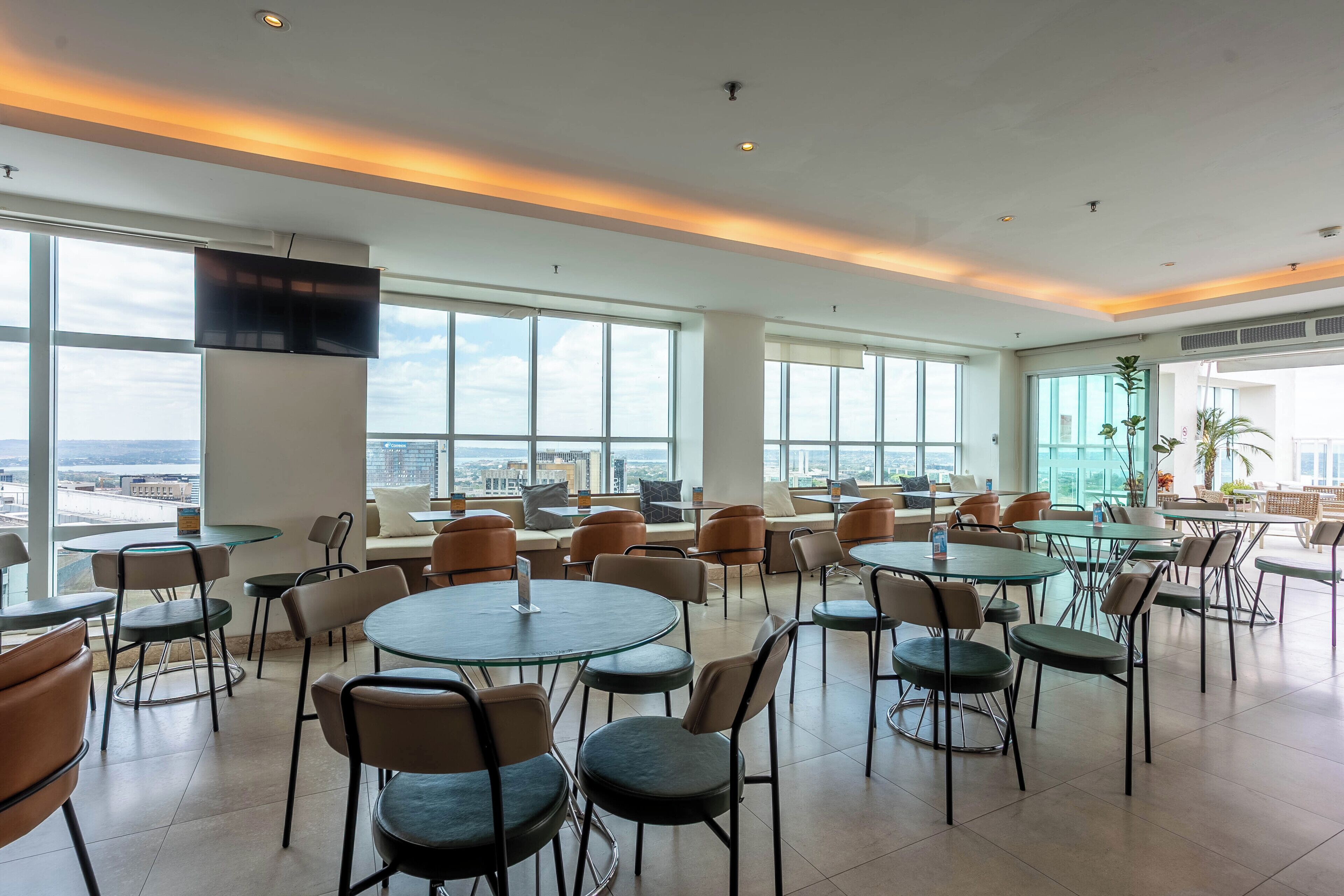 A spacious indoor area with multiple tables and chairs, large windows offering a city view, and a flat-screen TV mounted on the wall.