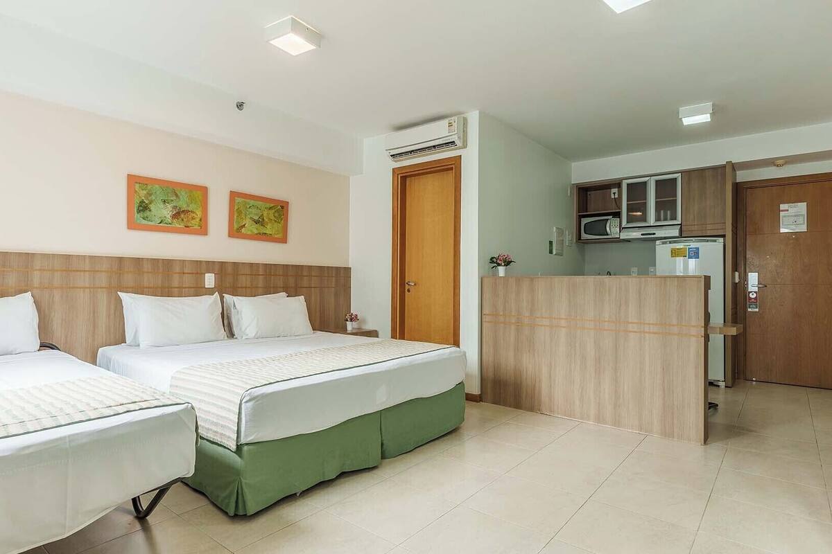 Quarto de hotel com duas camas, um espaço de cozinha compacto e uma porta que dá para outro cômodo.