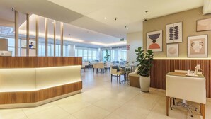 A modern reception area with a wooden reception desk, a large potted plant, and framed artwork on the walls. - Saint Moritz Hplus Express (Brasília)