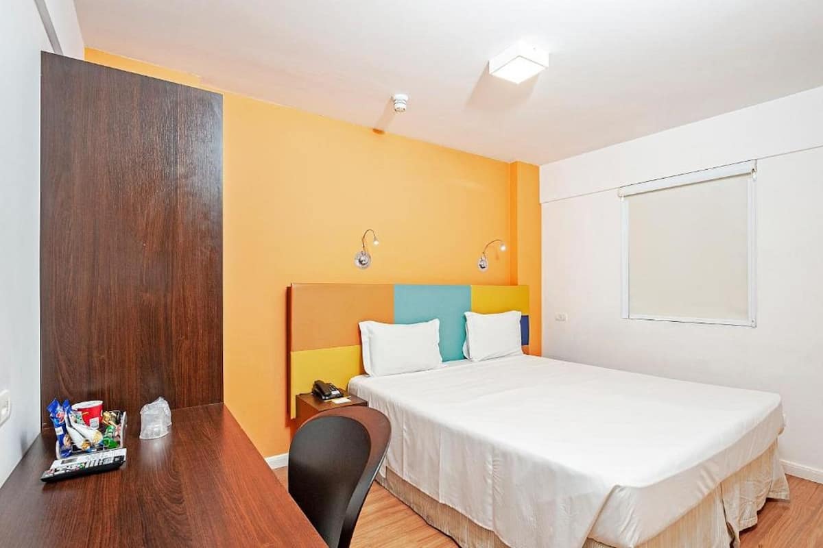 A hotel room with a bed, a desk, a chair, a nightstand, and a window.