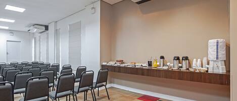 Un salón de conferencias con filas de sillas, un mostrador de buffet con comida y bebidas, y una unidad de aire acondicionado en el techo.
