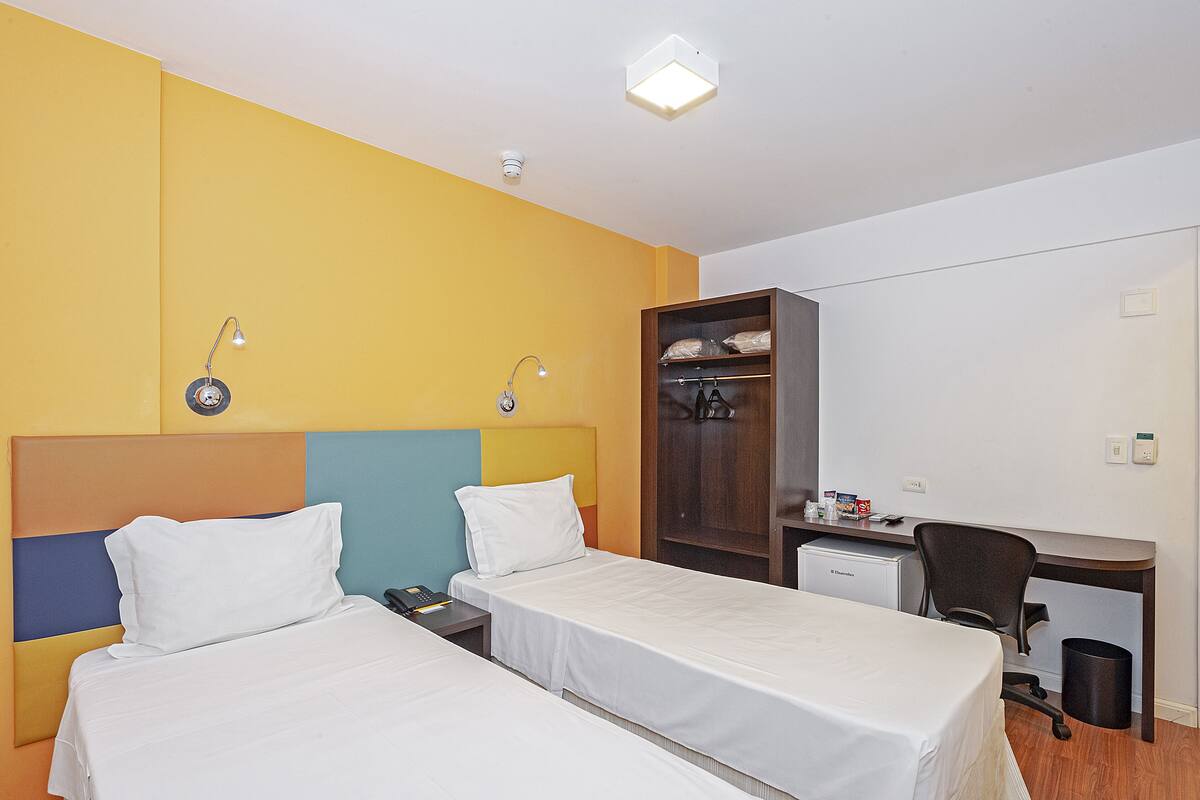 A hotel room with two beds, a desk, a chair, a wardrobe, and a small desk lamp.