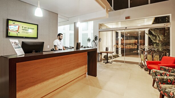 A reception desk with a monitor displaying a sign, a man standing behind it, and a seating area with a red chair and a plant. - Camboriú Express (Balneário Camboriú)