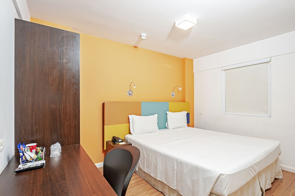 A hotel room with a bed, a desk, a chair, a nightstand, and a window.
