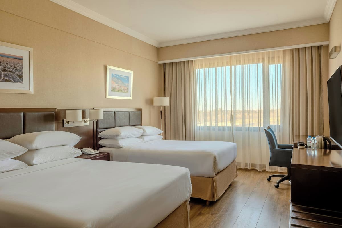 A hotel room with two beds, a desk, a chair, a TV, and a window with curtains.