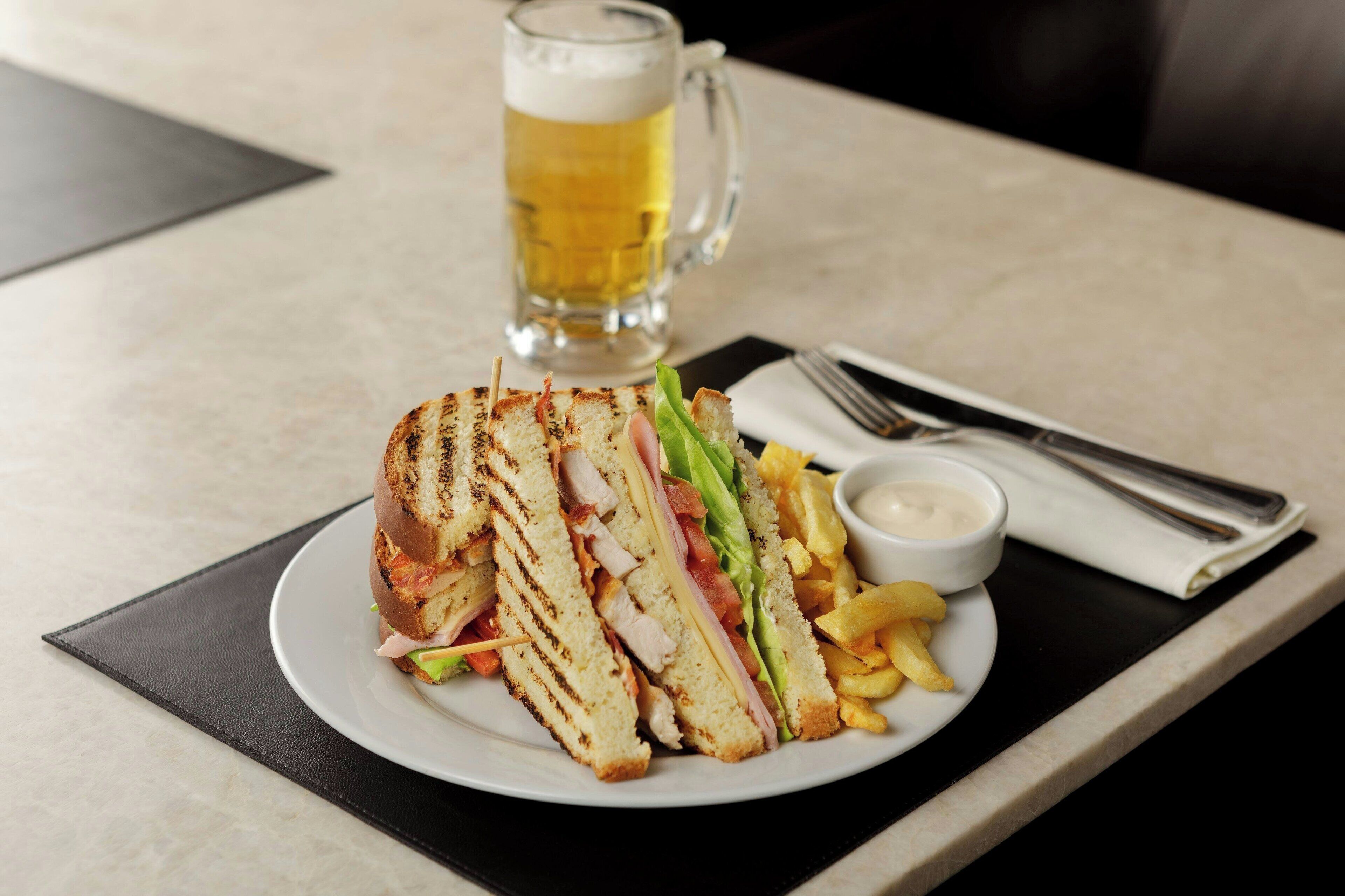 Ein Teller mit einem gegrillten Sandwich, Pommes und einer kleinen Schale Soße, dazu ein Glas Bier auf einem Tisch.