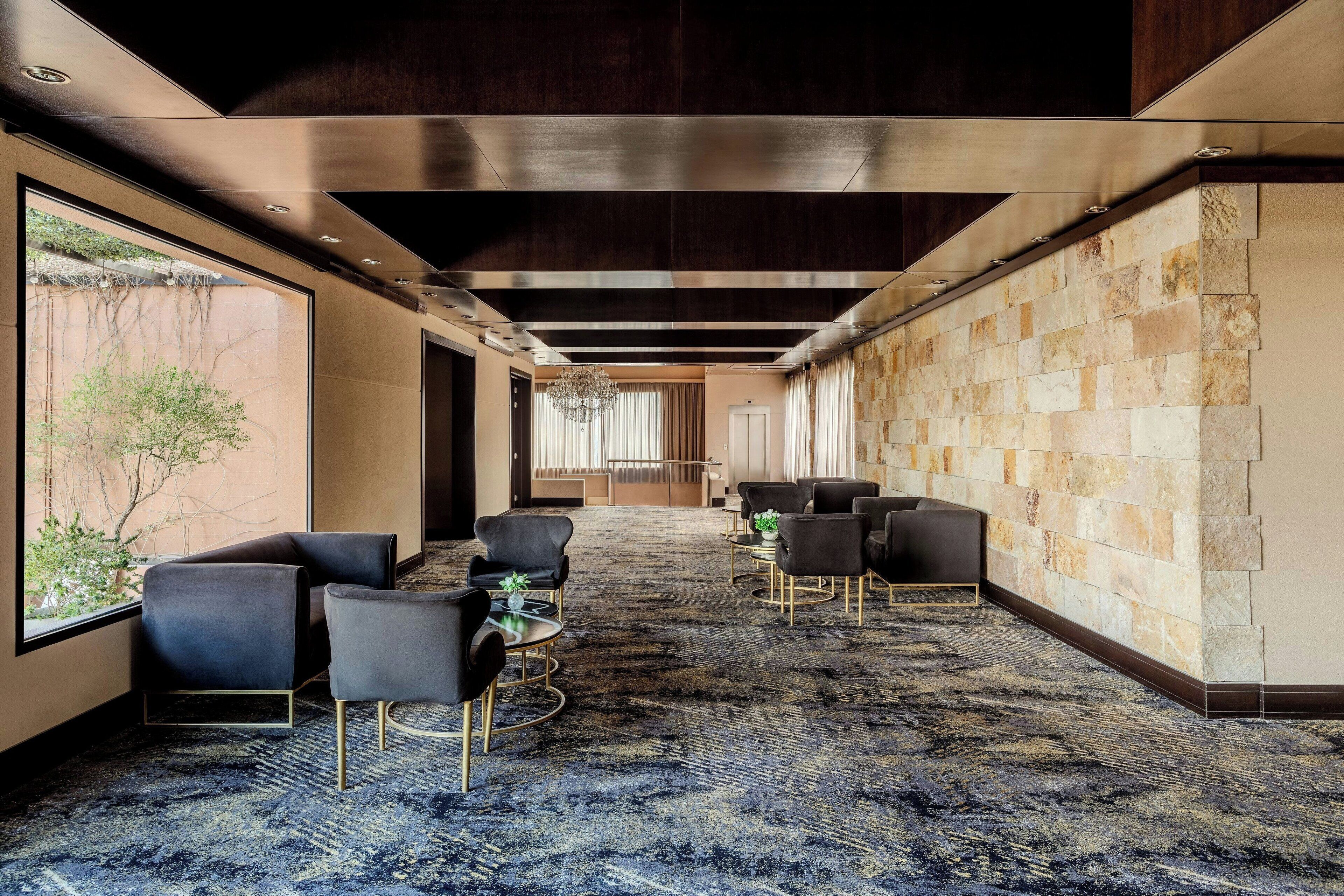 Un hall d’hôtel moderne avec un long couloir, un espace détente avec des chaises et des tables, un lustre et un mur en pierre.