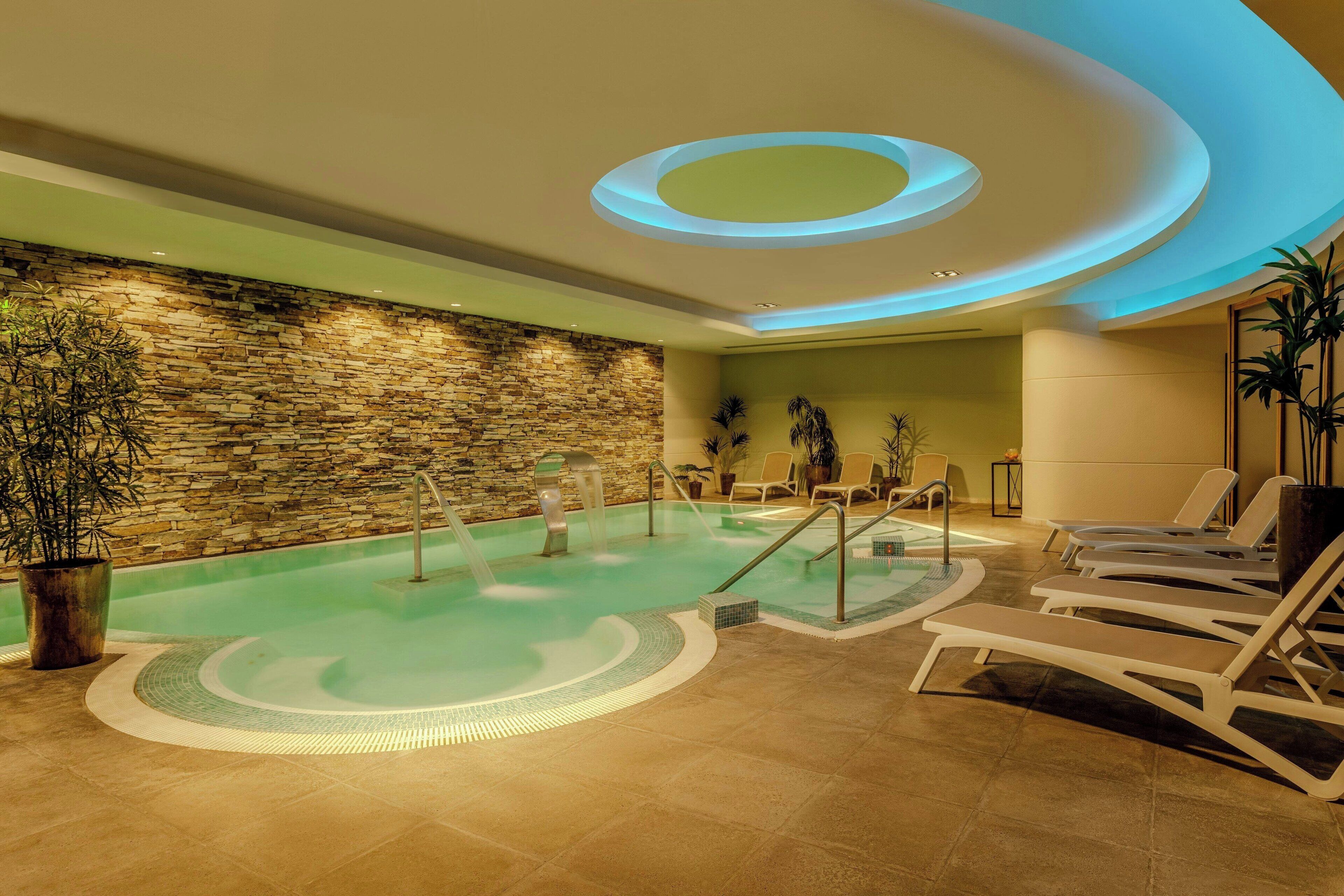 A spa area with a stone wall, a pool, lounge chairs, and a ceiling with circular lighting.