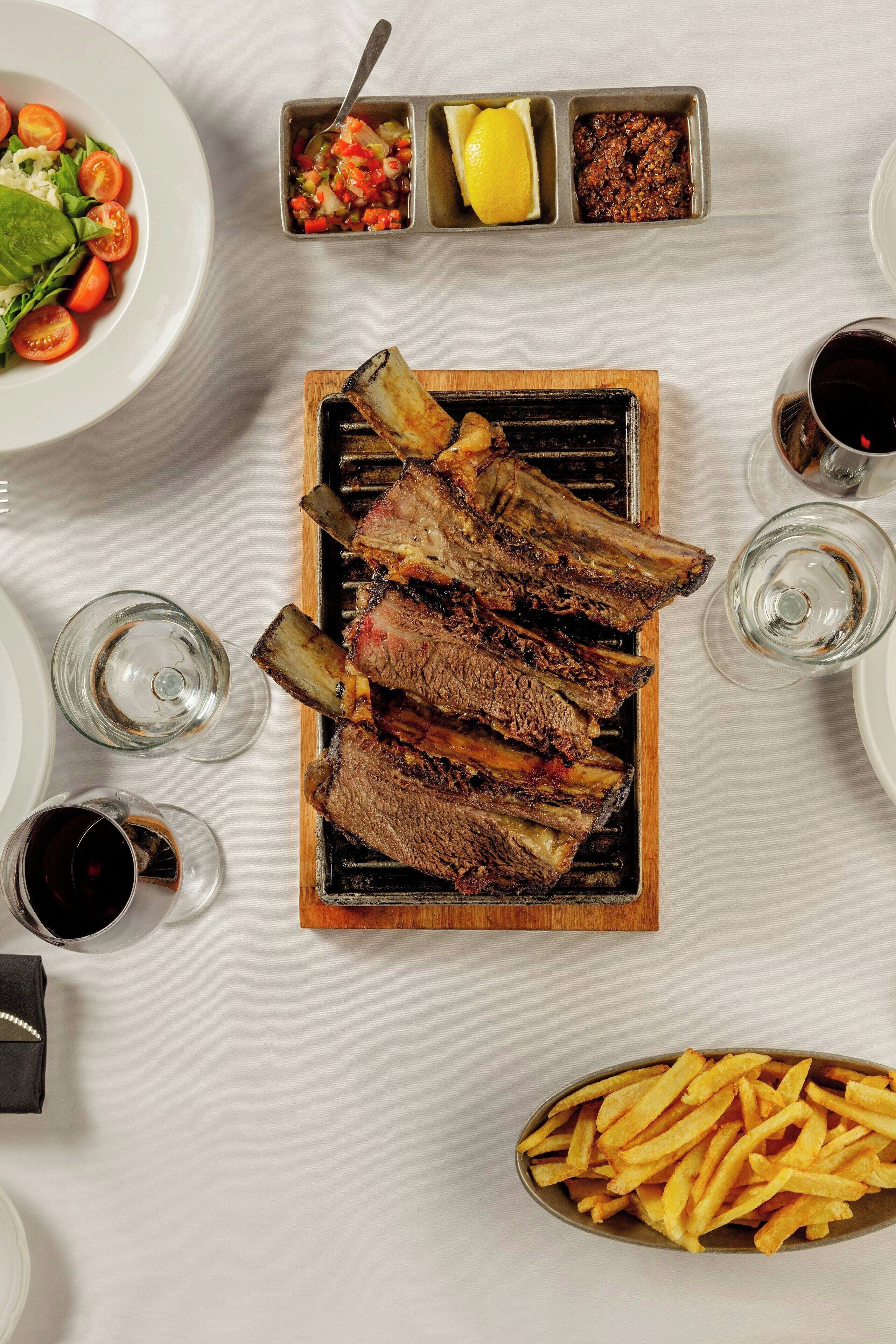 Una mesa con costillas a la parrilla, una ensalada, un recipiente con salsas, una copa de vino y una porción de papas fritas.