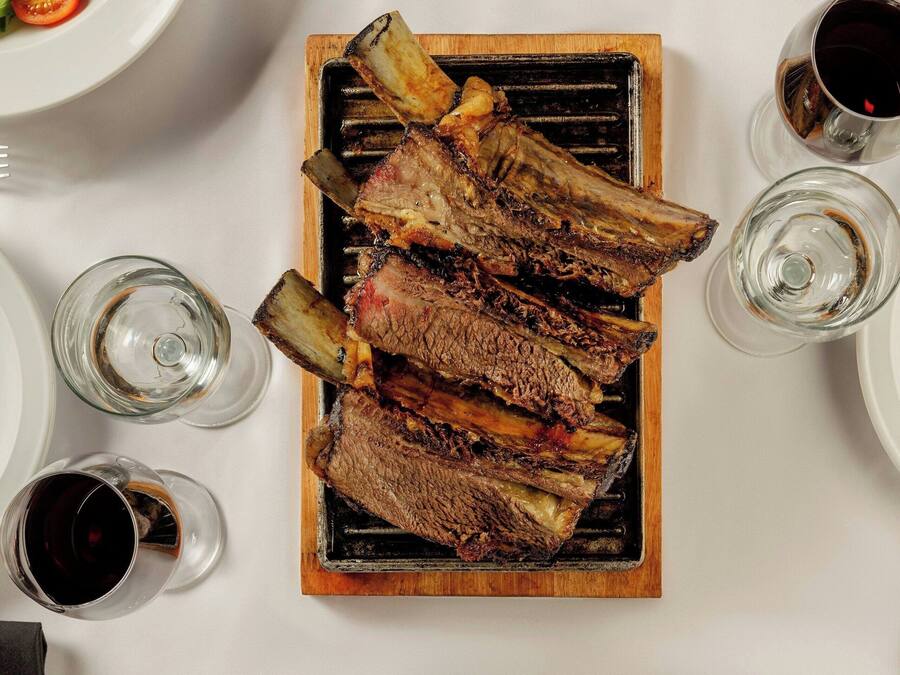 Una mesa con costillas a la parrilla, una ensalada, un recipiente con salsas, una copa de vino y una porción de papas fritas.