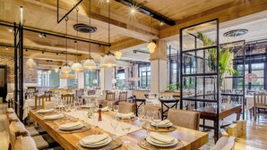 Àmplia zona de dinar amb taules de fusta llargues preparades per a diversos convidats, amb il·luminació penjant moderna i grans finestres que ofereixen vistes a l'exterior.