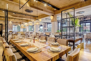 A spacious dining area with long wooden tables set for multiple guests, featuring modern pendant lighting and large windows offering views of the outdoors. - Marriott Hotel Buenos Aires Ezeiza Airport (Ciudad Evita)