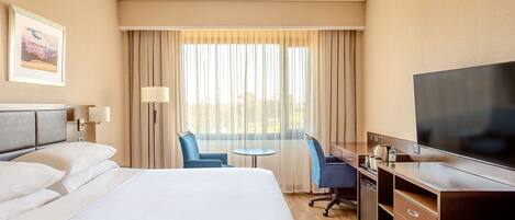 Habitación de hotel con una cama grande, un escritorio con silla, televisión y ventana con cortinas.