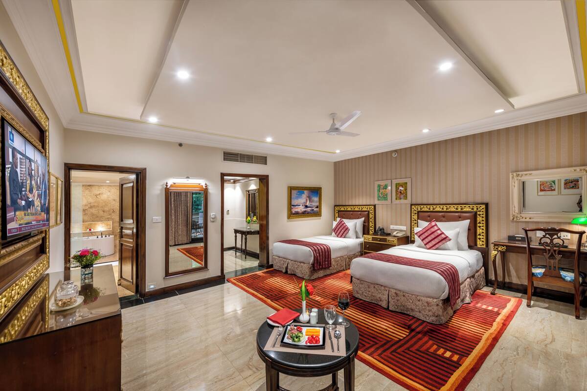 A hotel room with two beds, a desk, a chair, a TV, and a dining area with a table set for two.