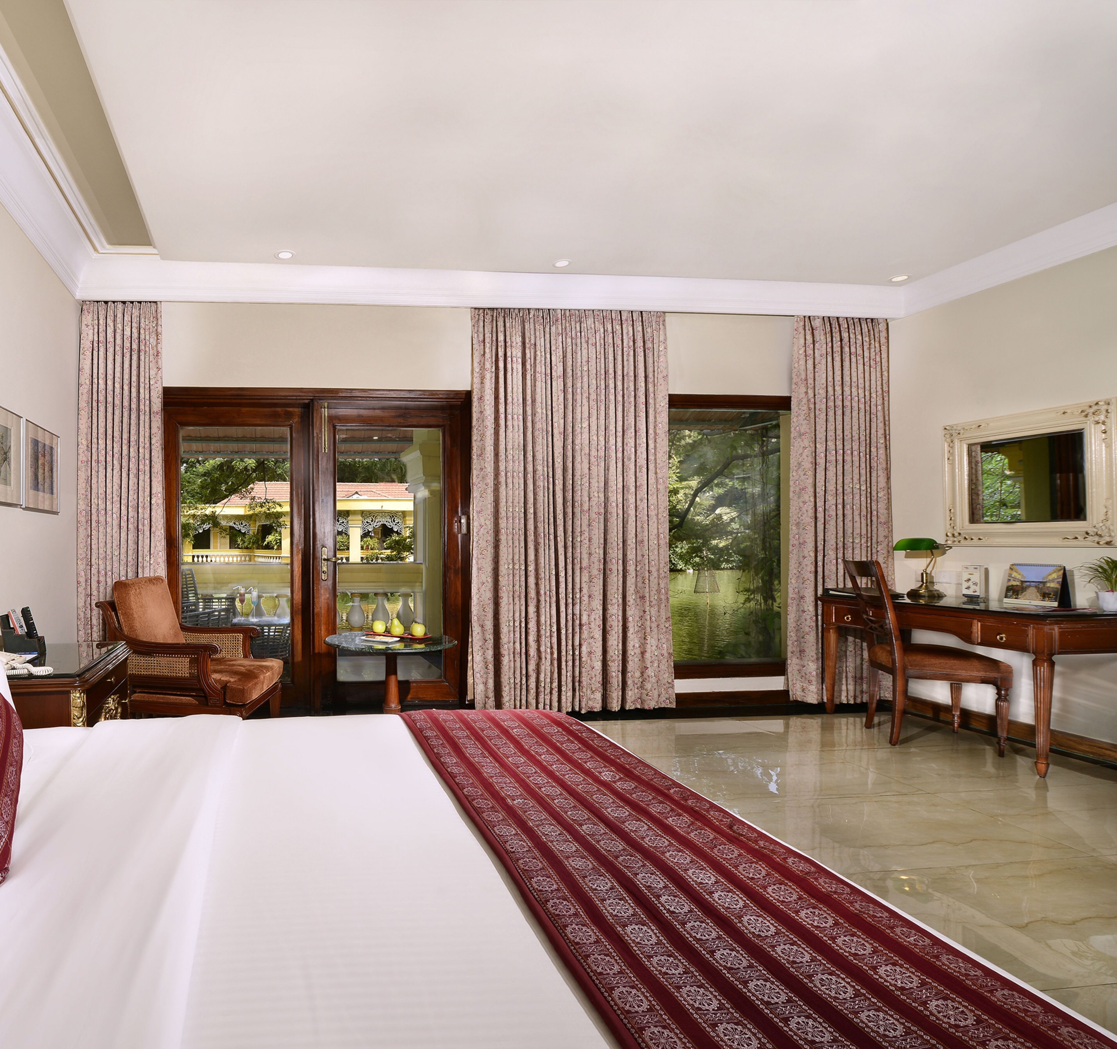 A hotel room with a bed, a desk, a chair, a table with fruit, and a view of greenery outside.
