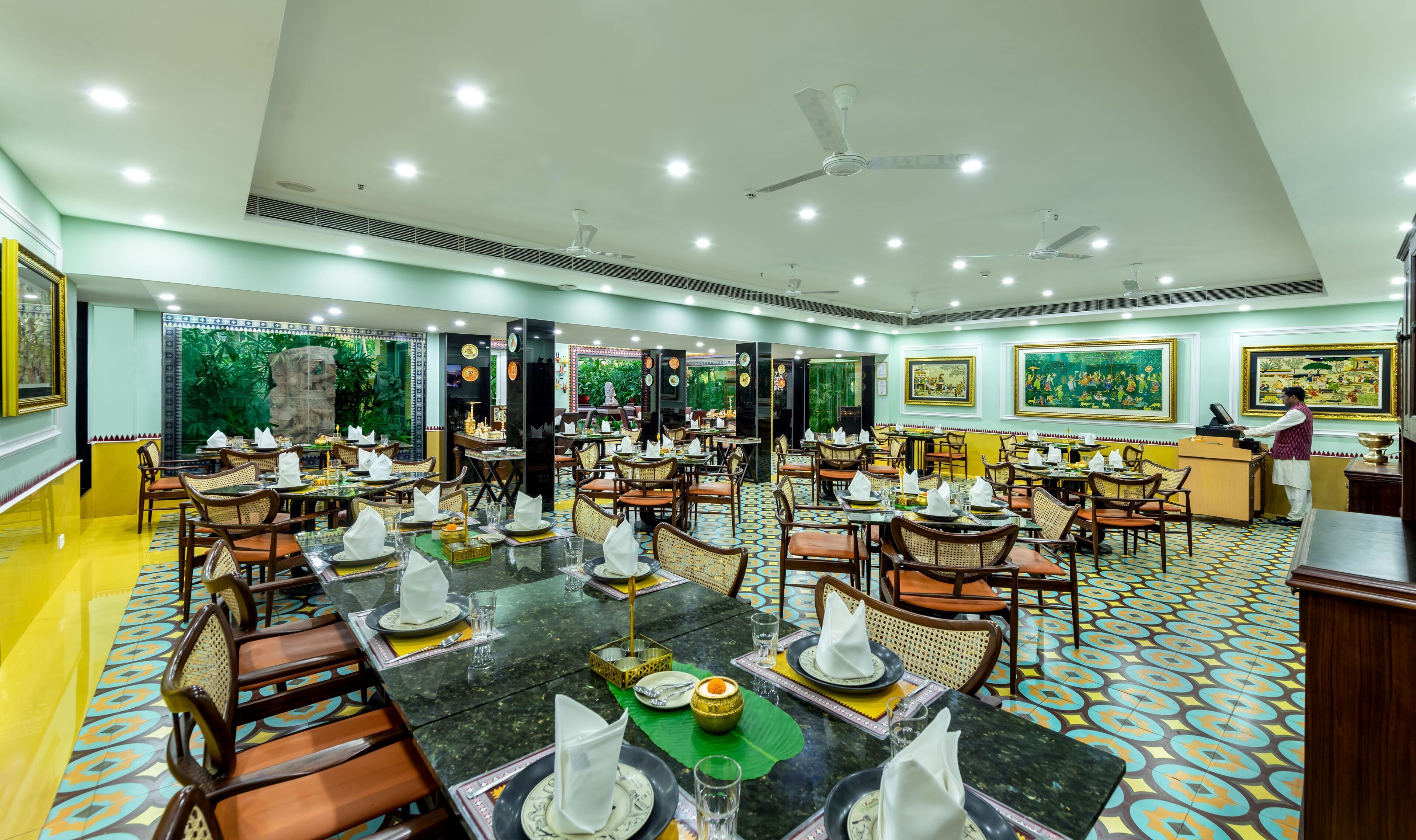 A spacious restaurant with multiple dining tables, ceiling fans, and decorative wall paintings.