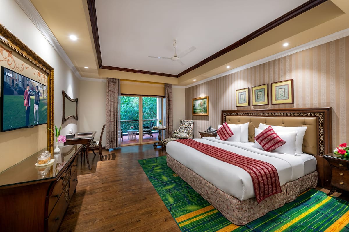 A spacious bedroom with a large bed, a desk, and a chair. There is a window with a view of greenery outside.