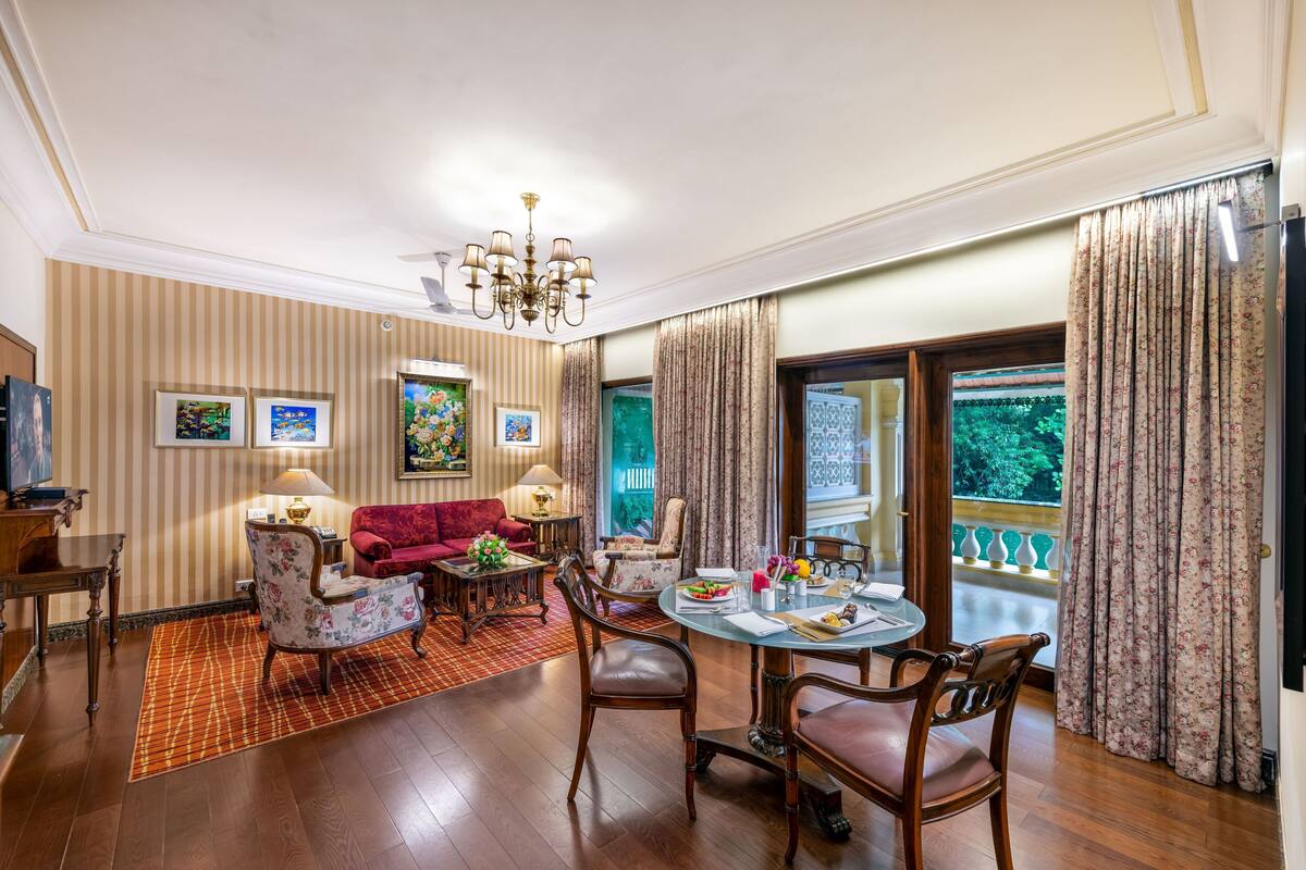 A dining area with a round table, chairs, and a red sofa. There is a chandelier above the table and a window with curtains.